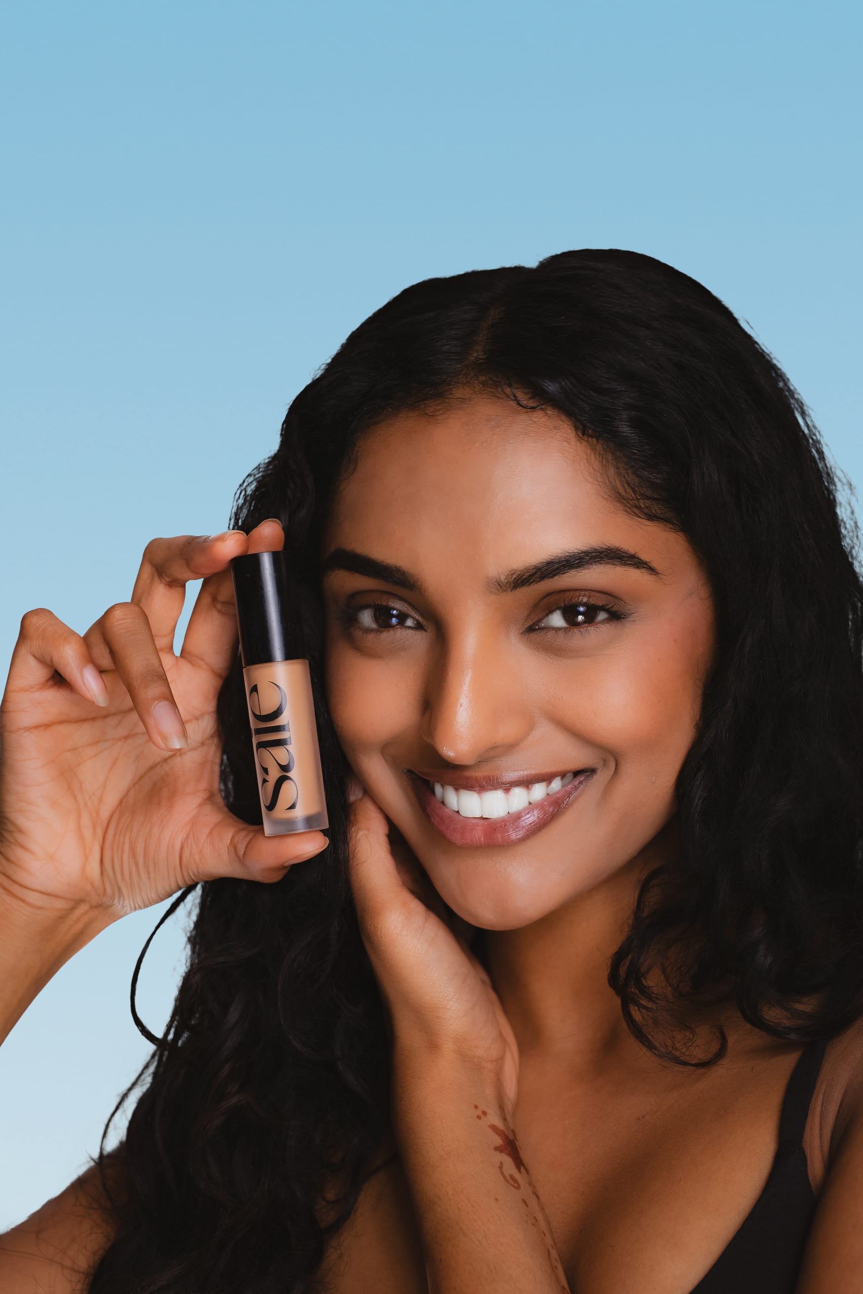 Smiling beauty test shot holding a Saie concealer, blue background