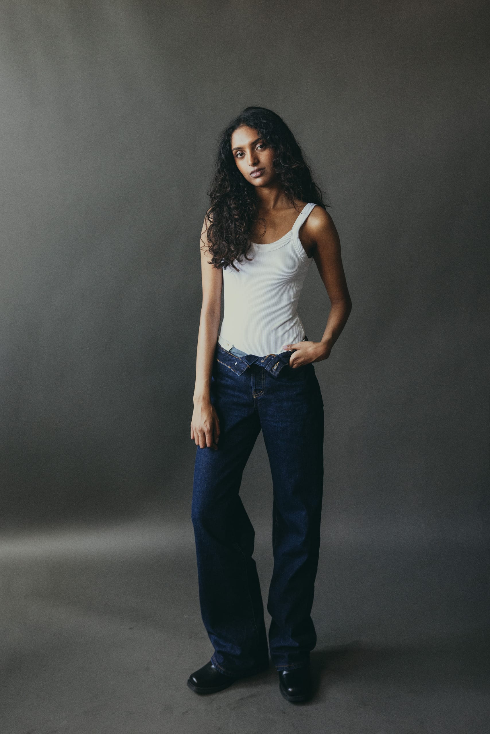 Full length in a white tank and dark denim against a grey studio backdrop