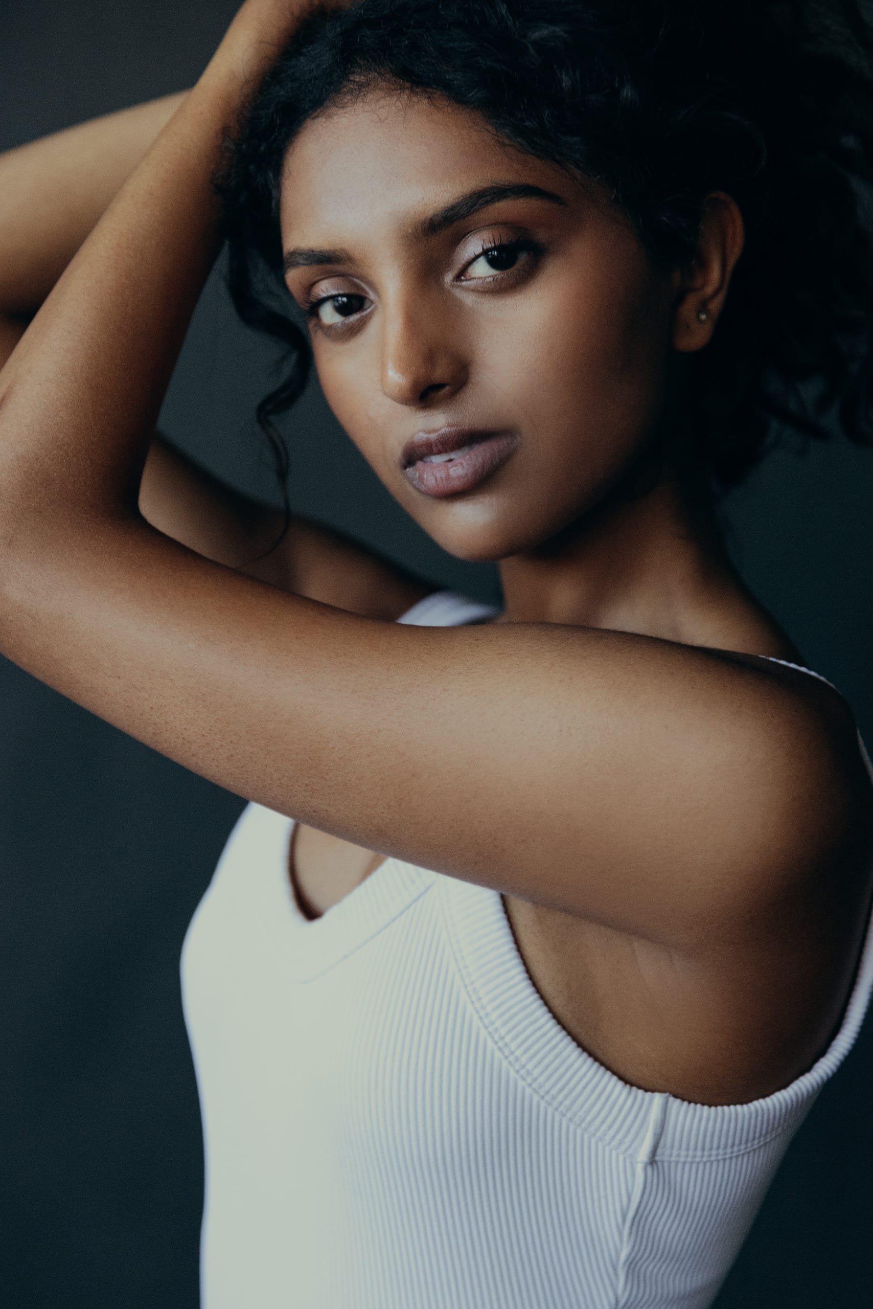 Portrait in a white tank with one arm raised, grey studio backdrop