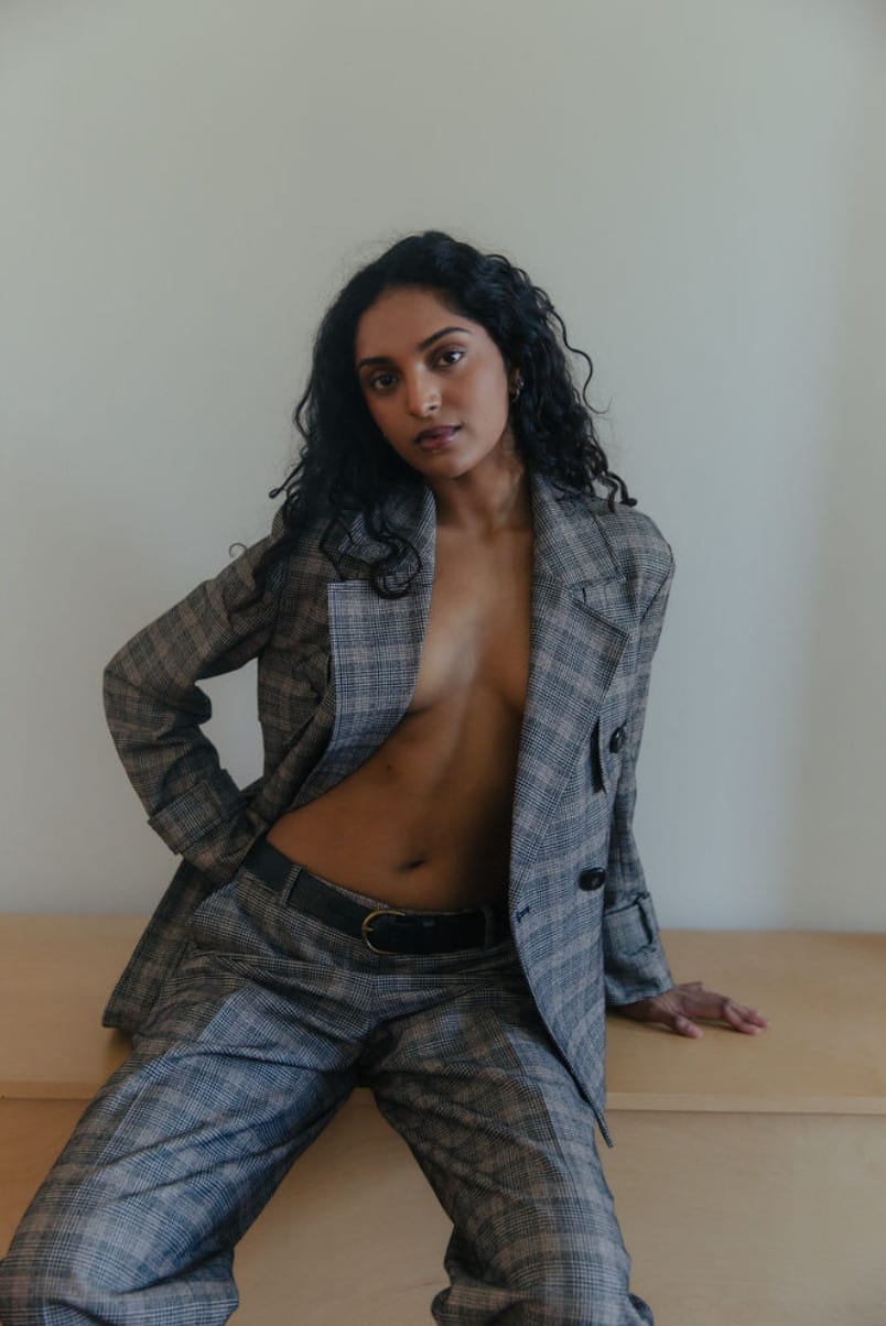 Editorial test in an open grey plaid suit, seated against a pale wall