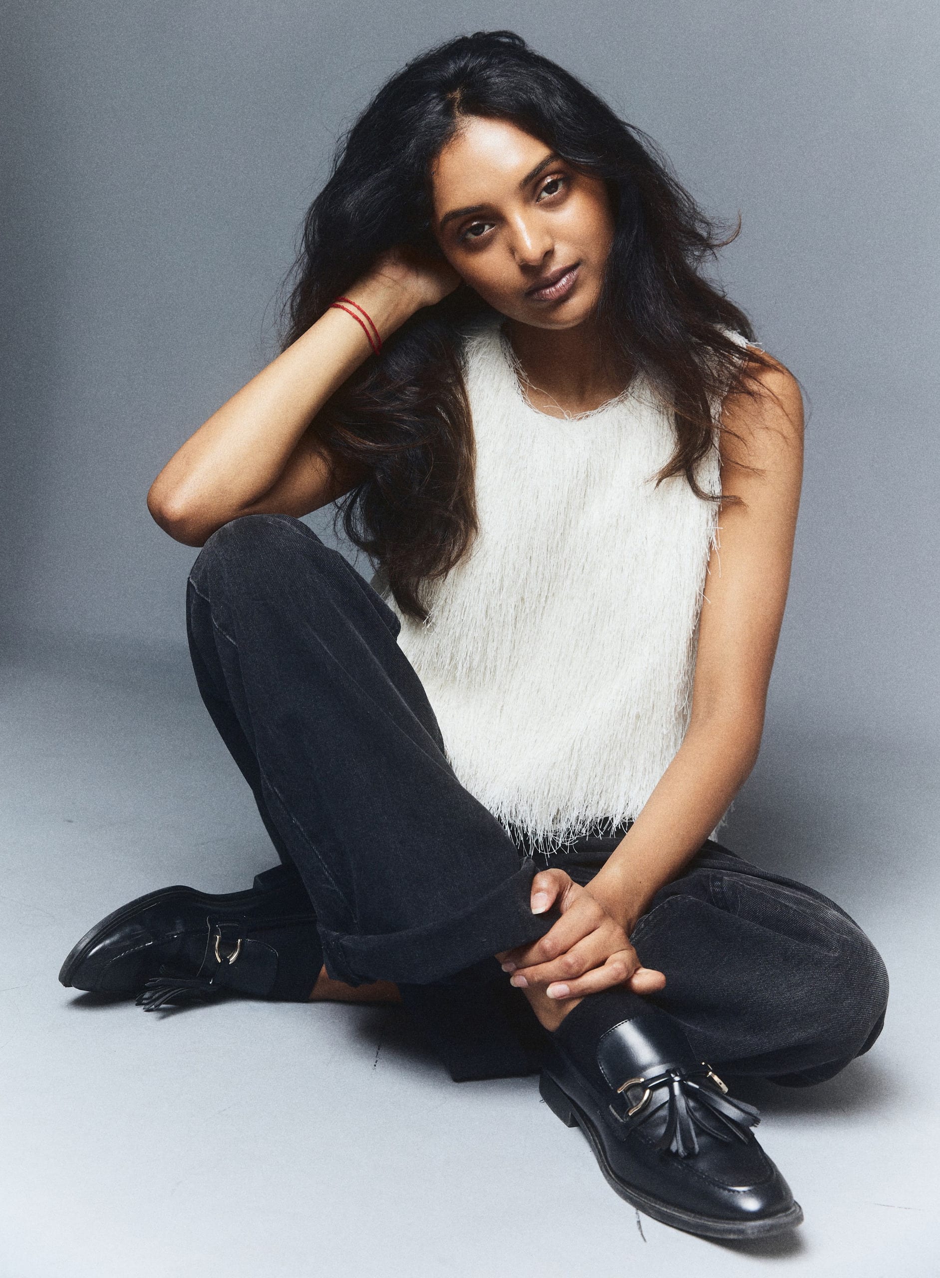 Seated on the floor in a white knit top and dark denim, grey studio backdrop