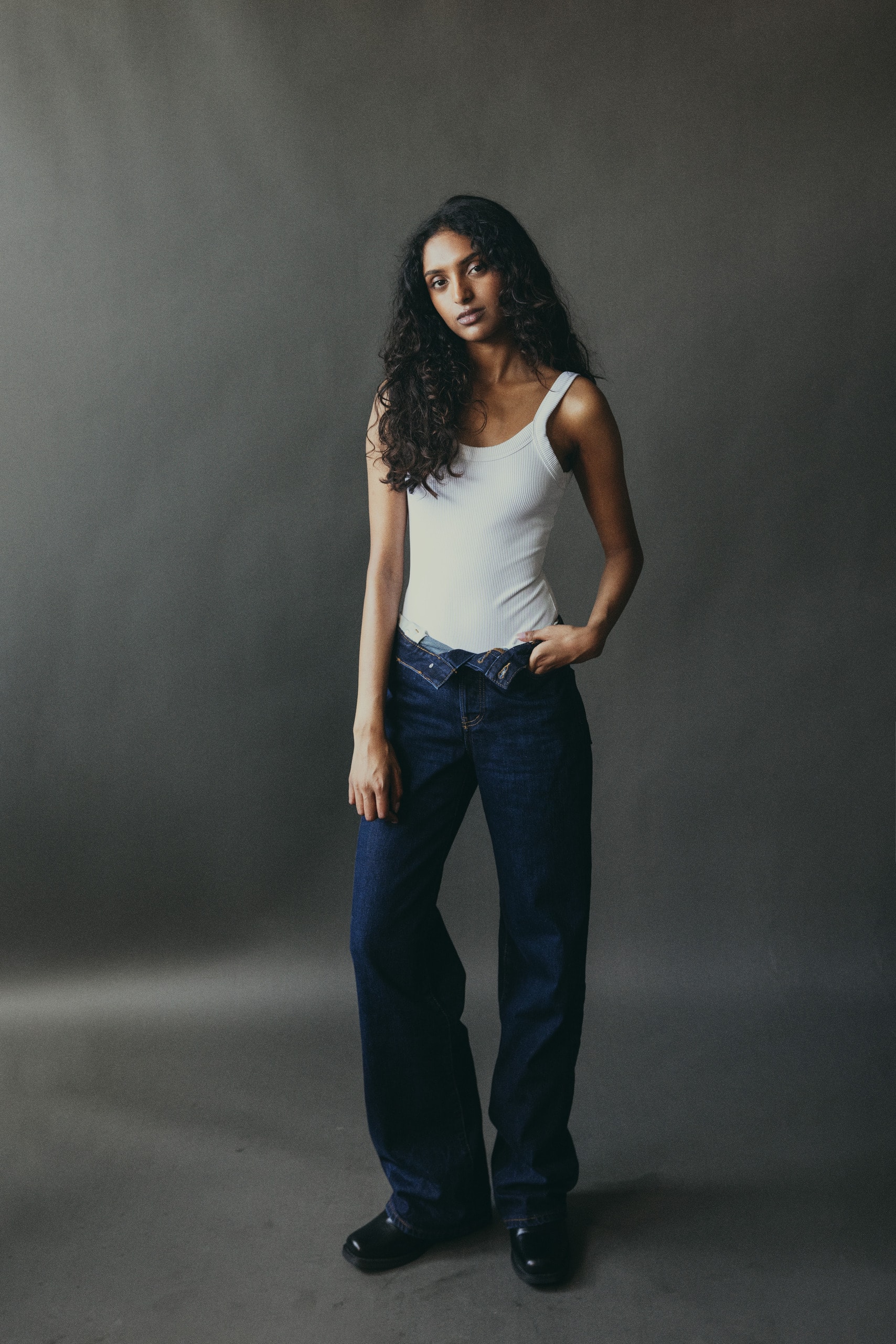 Full length in a white tank and dark denim against a grey studio backdrop