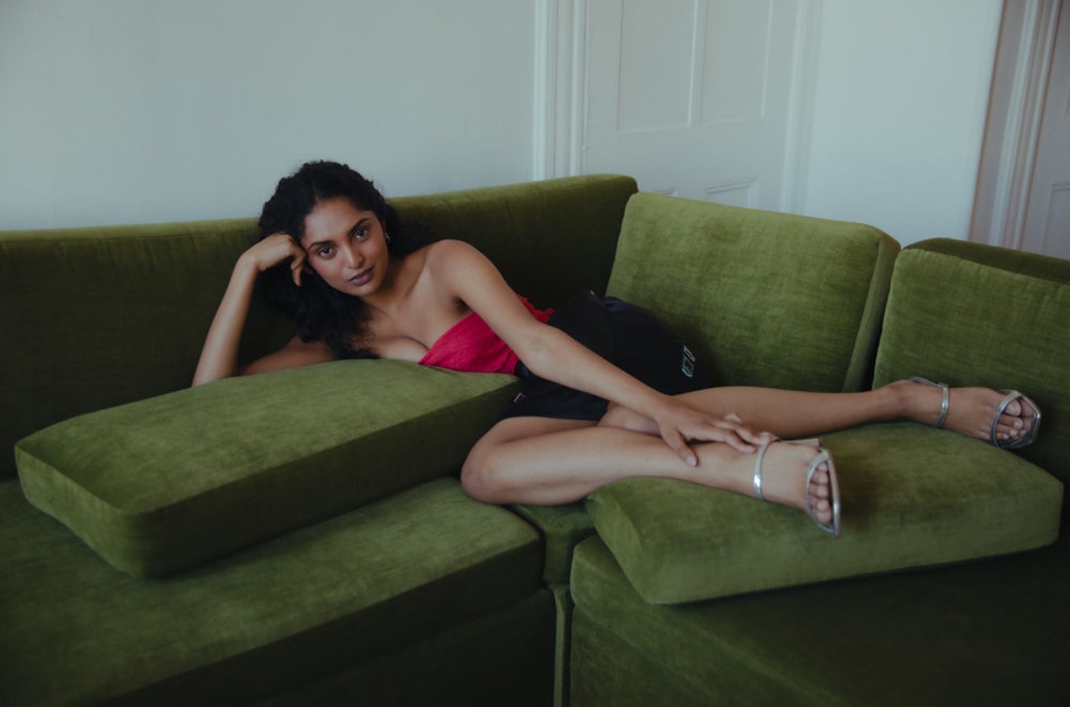 Editorial test reclining on a green velvet sofa in a red top and silver heels